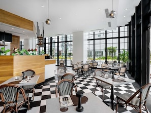 Un restaurante moderno con suelo a cuadros, sillas de ratán blanco y negro, y grandes ventanales con vistas al exterior.