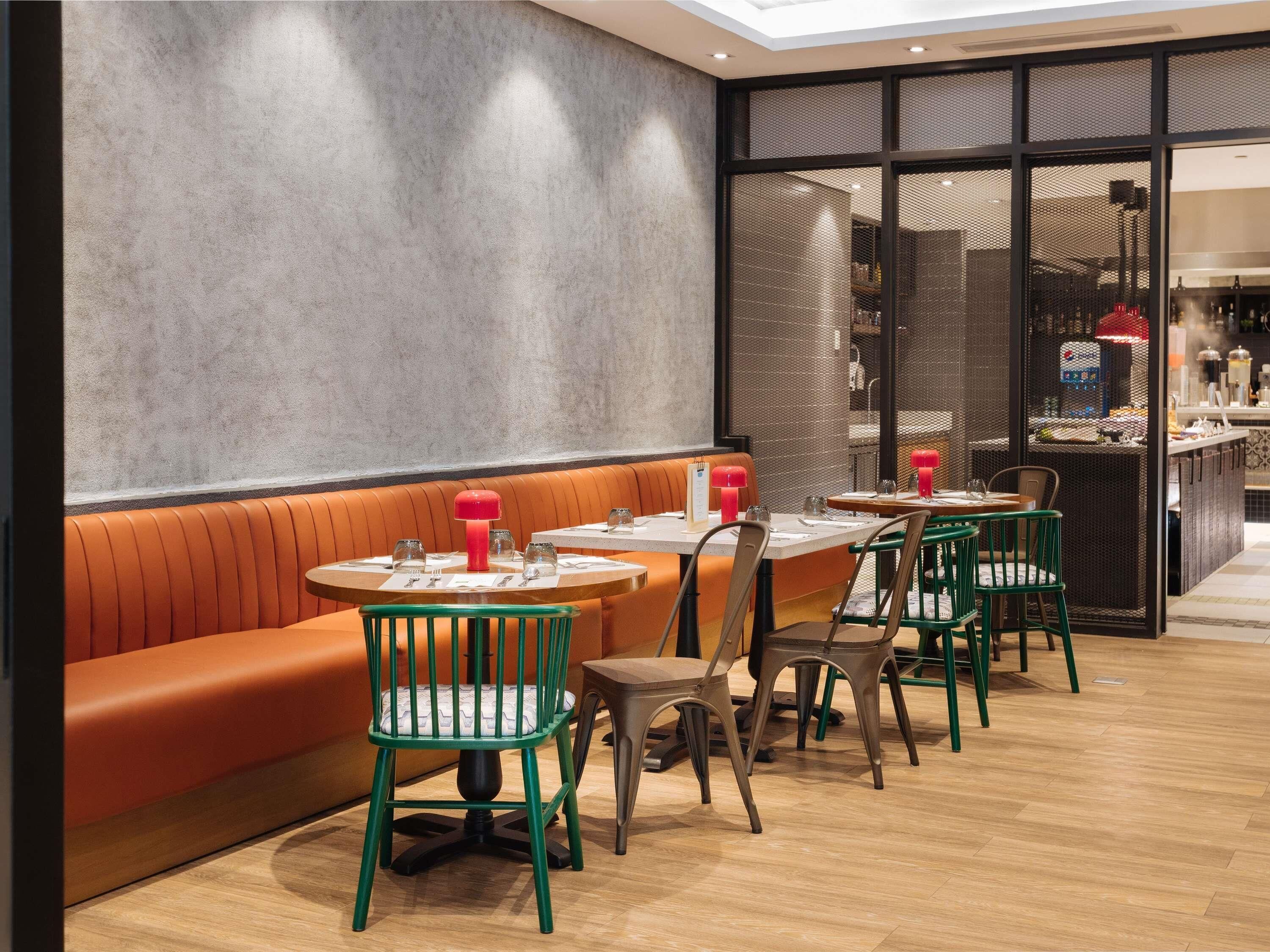 A modern restaurant interior with a long orange bench, round tables, and green chairs.
