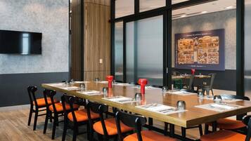 A conference room with a long table, orange chairs, a mounted TV, and a large window with a cityscape view.