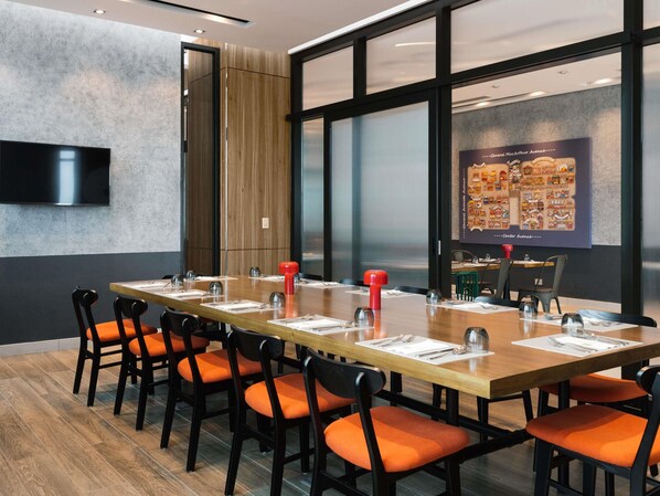 A conference room with a long table, orange chairs, a mounted TV, and a large window with a cityscape view.