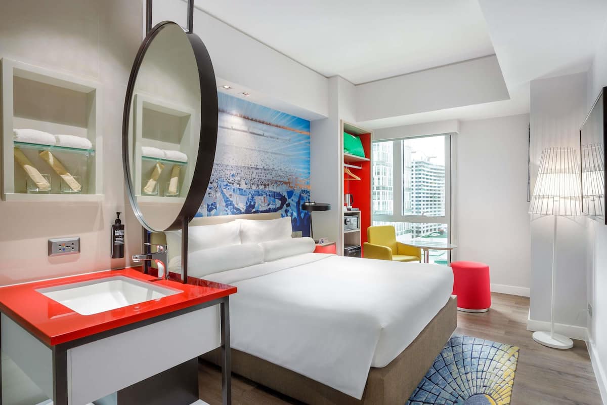 Ein modernes Hotelzimmer mit einem großen Bett, einem Badezimmer in Rot und Weiß, einem großen Spiegel und Blick auf die Stadt.