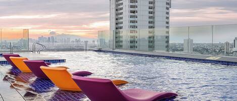 Une piscine sur le toit, agrémentée de chaises longues violettes et orange, d’une balustrade en verre et offrant une vue sur le panorama urbain en arrière-plan.