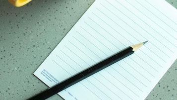 A notepad with lined paper, a pencil, and a yellow mug on a speckled surface.