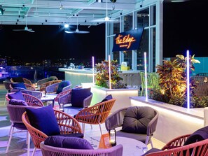 Een buitengebied met een bar, rieten stoelen en een uitzicht over de stad 's nachts.