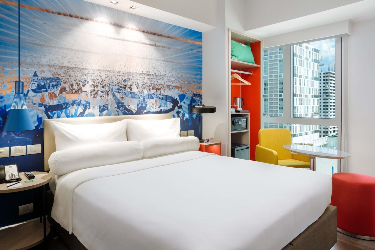 A modern hotel room with a large bed, a small desk, a chair, a red ottoman, and a city view through the window.