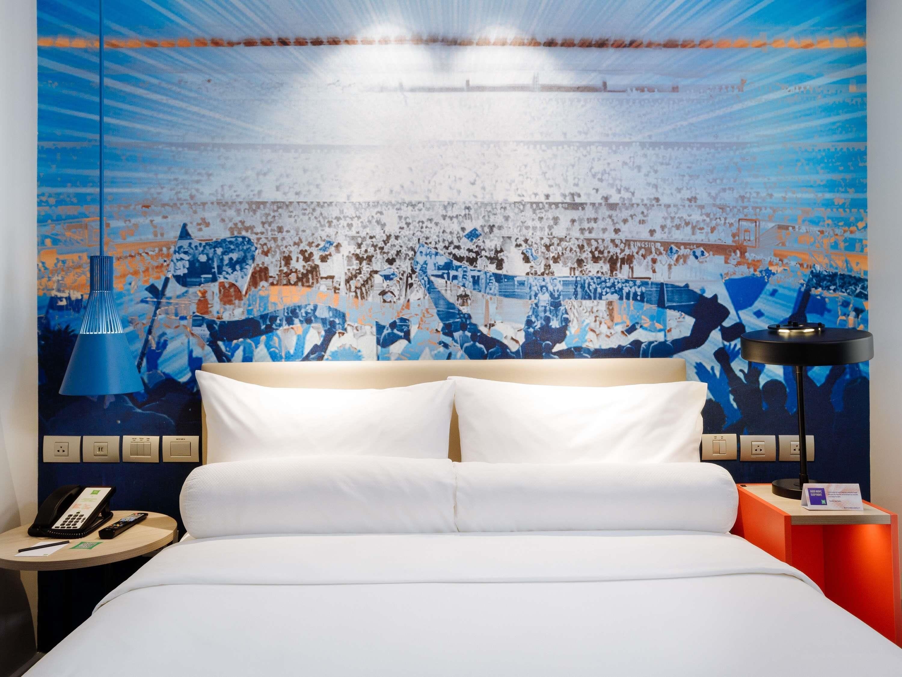 A hotel room with a bed, a nightstand with a phone and remote, a lamp, and a wall with a crowd scene.