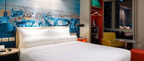 Habitación de hotel con cama, escritorio, silla, mesita, ventana con vista a la ciudad y una obra de arte abstracta y colorida en la pared.