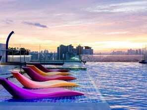 A rooftop infinity pool with colorful lounge chairs, a city skyline in the background, and a sunset. - Ibis Styles Manila Araneta City (Quezon City)