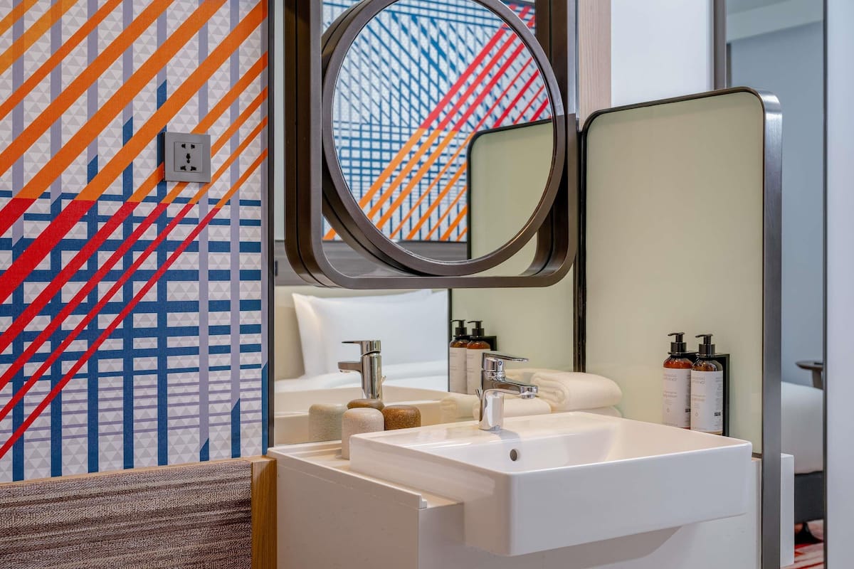Une salle de bain moderne avec un lavabo blanc, deux robinets, un miroir et un mur décoré d’un motif.