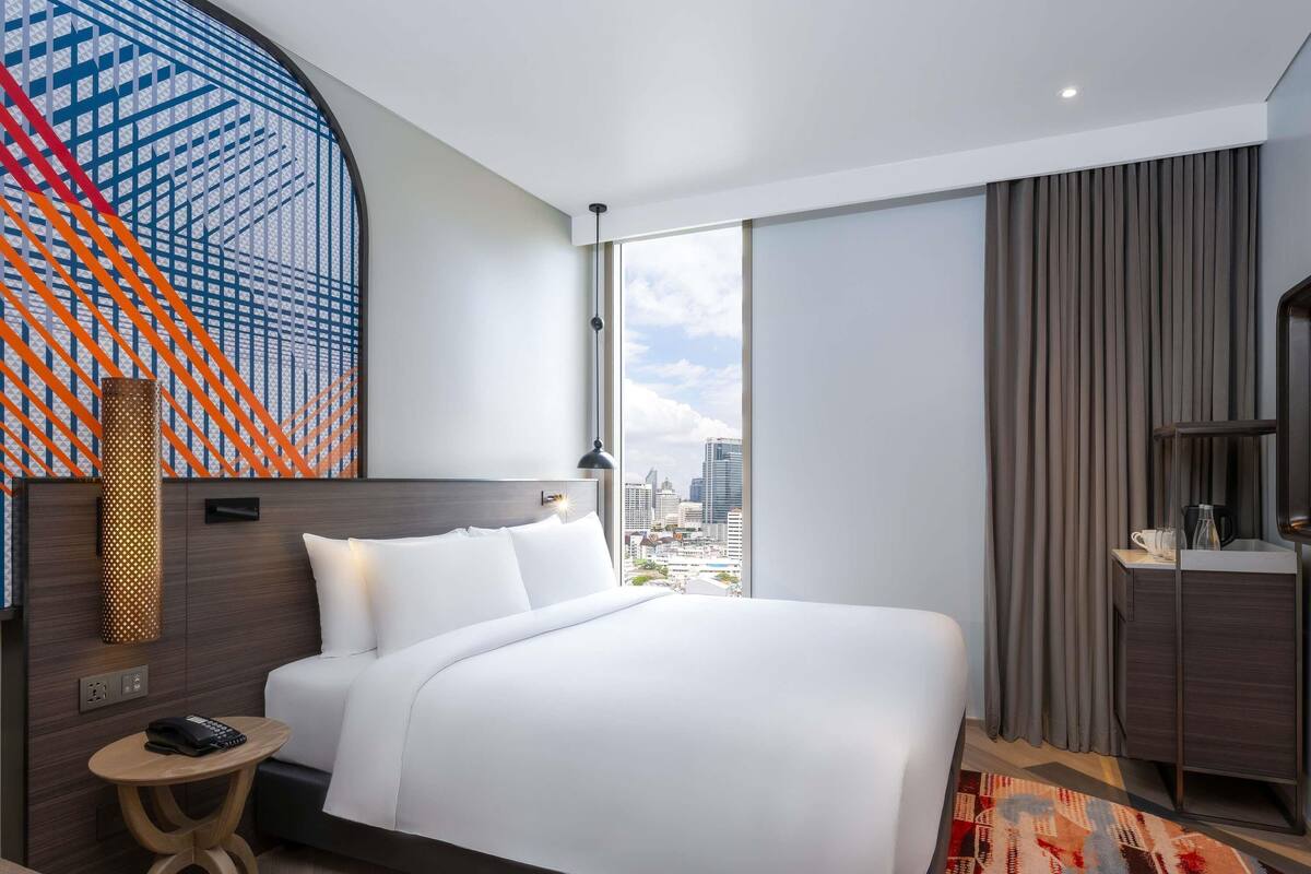 Une chambre d’hôtel moderne avec un grand lit, une petite table de chevet, un bureau avec une lampe et une vue sur le panorama urbain.