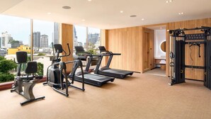 A gym with treadmills, elliptical machines, and weightlifting equipment, large windows with a city view, and wooden panel walls. - Mercure Bangkok Surawong (Bangkok)