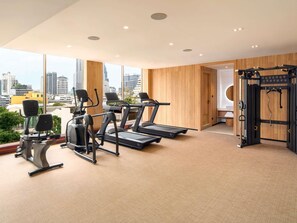 A gym with treadmills, elliptical machines, and weightlifting equipment, large windows with a city view, and wooden panel walls. - Mercure Bangkok Surawong (Bangkok)