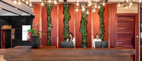 A reception desk with a wooden finish, two receptionists, and a wall with vertical wooden slats and greenery.