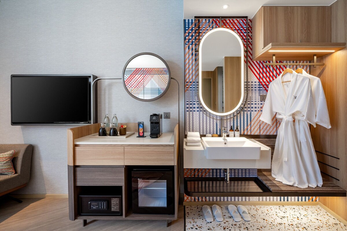 Een moderne hotelkamer met een flatscreen televisie, een koffiezetapparaat, een magnetron, een wastafel met spiegel, en een badjas hangend bij de deur.