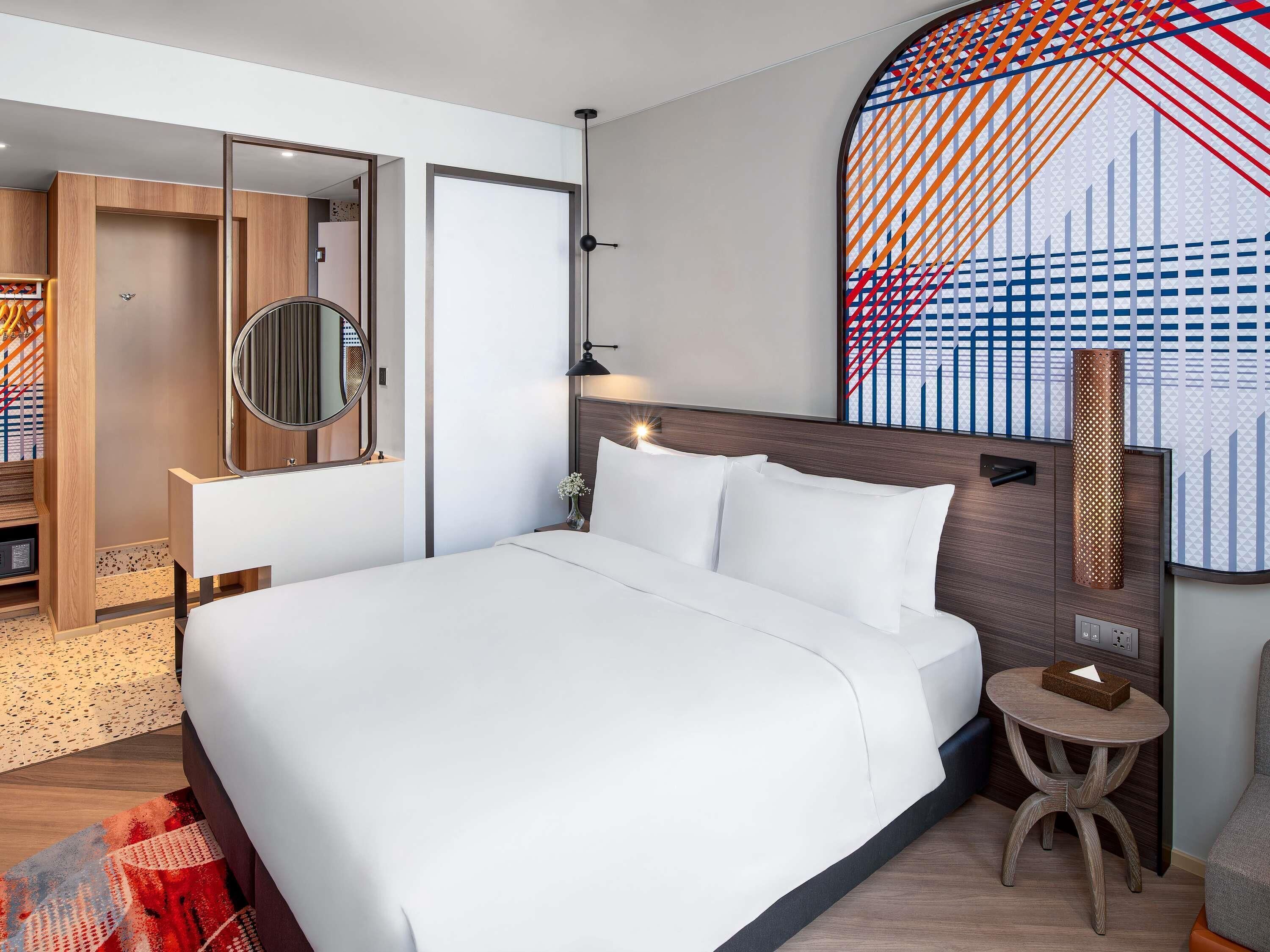 A modern hotel room with a large bed, a round mirror, a small round table, and a unique wall art piece.