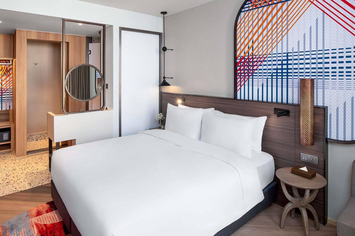 Une chambre d’hôtel moderne avec un grand lit, un miroir rond, une petite table ronde et une œuvre d’art murale originale.