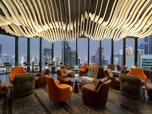 Un salon moderne avec des fauteuils en cuir, un plafond haut doté d’un éclairage original et une vue sur la ville.