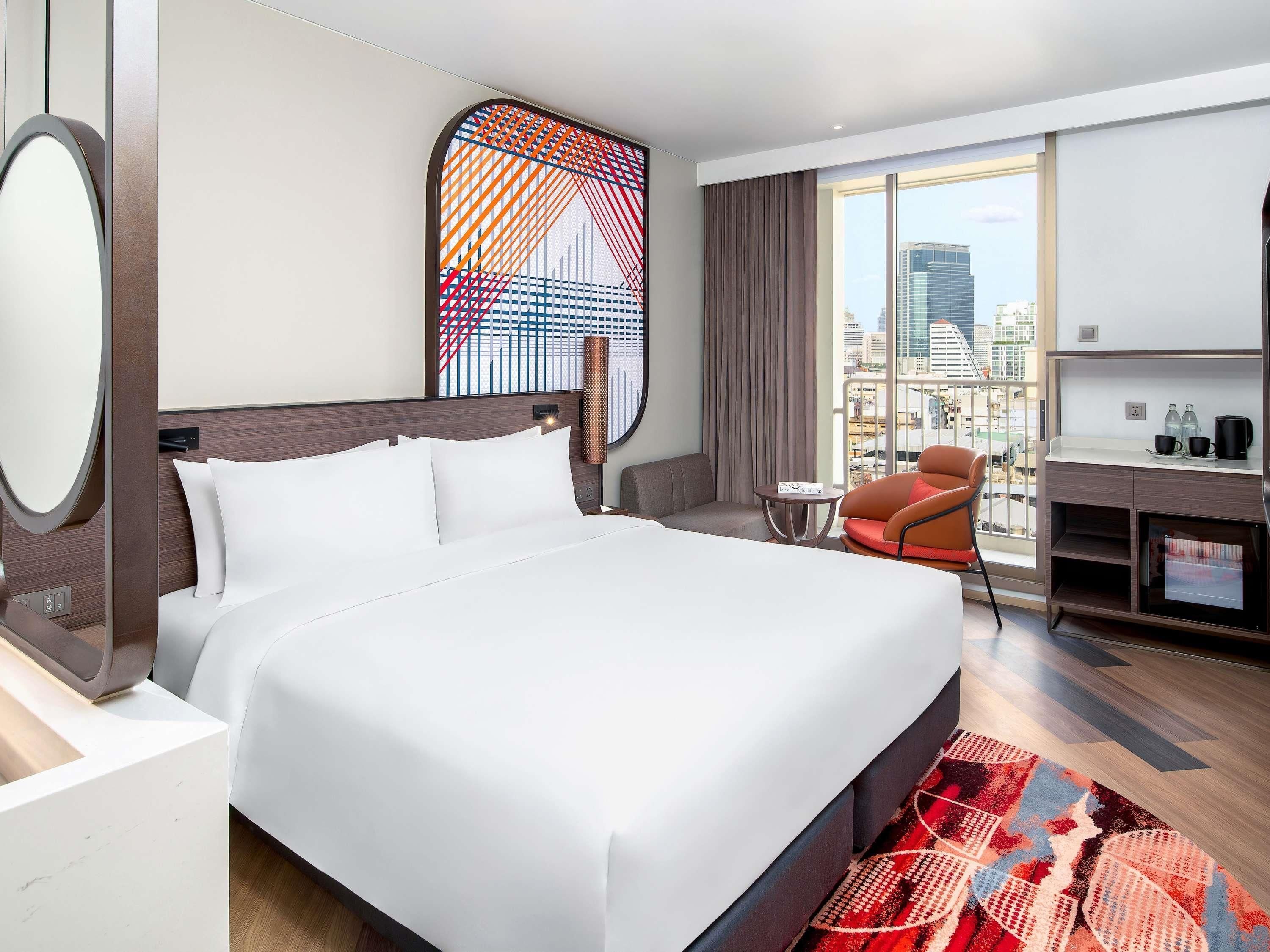 A modern hotel room with a large bed, a desk, a chair, and a view of the city.