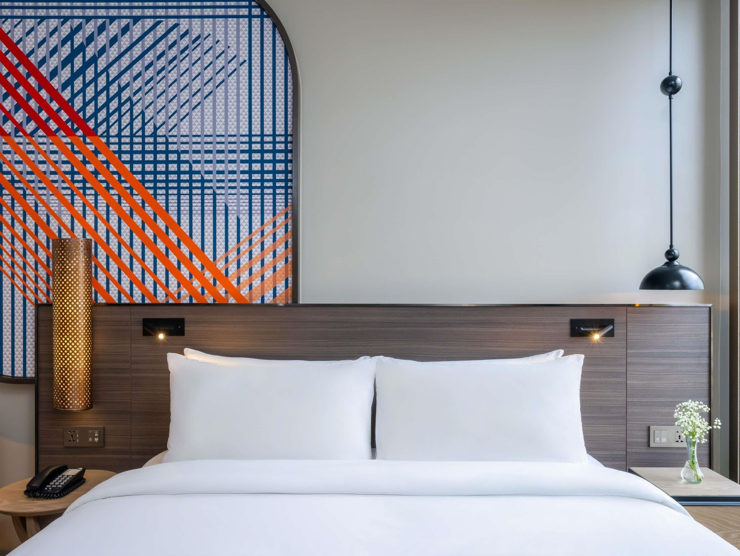 Ein Hotelzimmer mit einem ordentlich bezogenen Bett, einer Wandlampe, einem Telefon auf einem Nachttisch und einer dekorativen Vase mit Blumen.