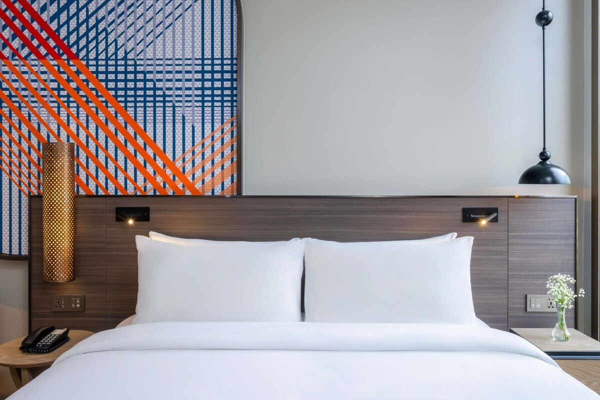 Une chambre d’hôtel avec un lit soigneusement fait, une lampe fixée au mur, un téléphone sur une table de chevet et un vase décoratif avec des fleurs.