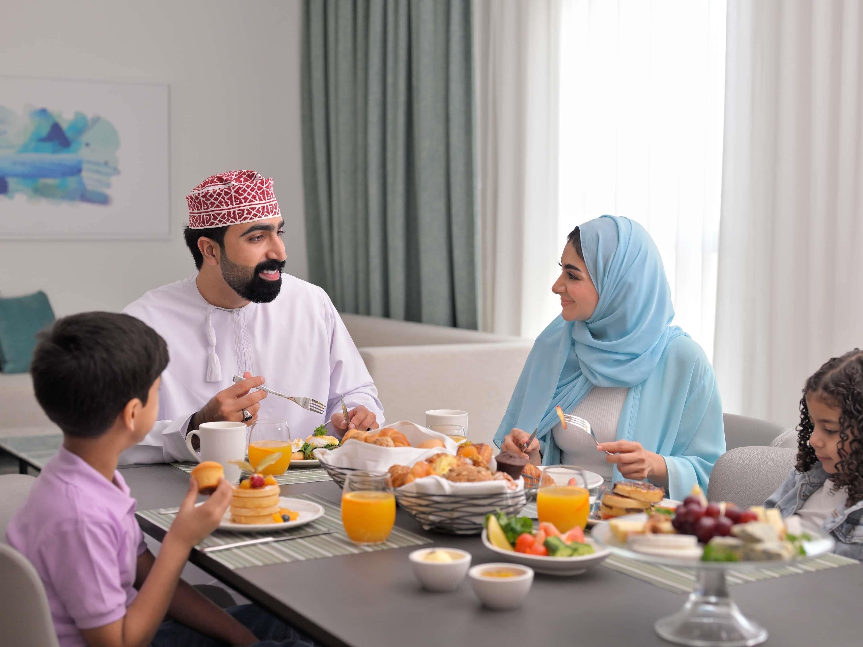 A family dining at a table with breakfast items, a child, and a woman in traditional attire.