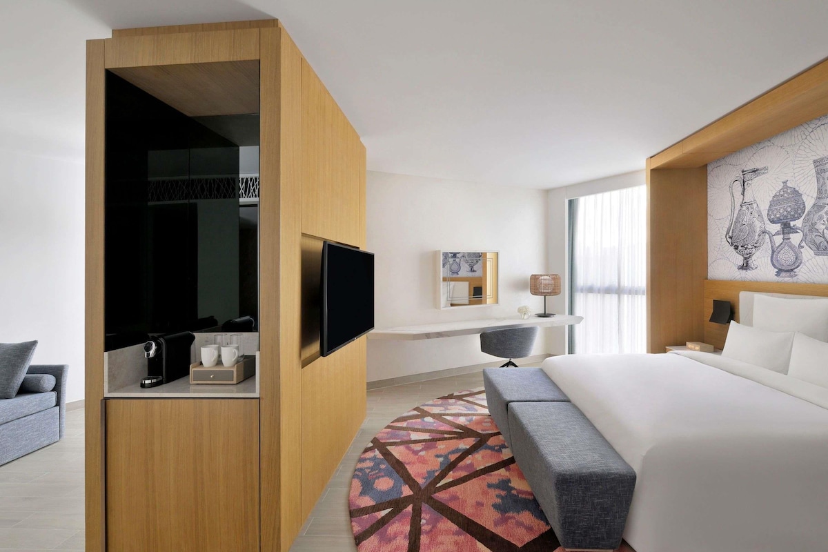 Habitación de hotel con cama, televisión montada en un panel de madera, escritorio con silla y alfombra estampada.