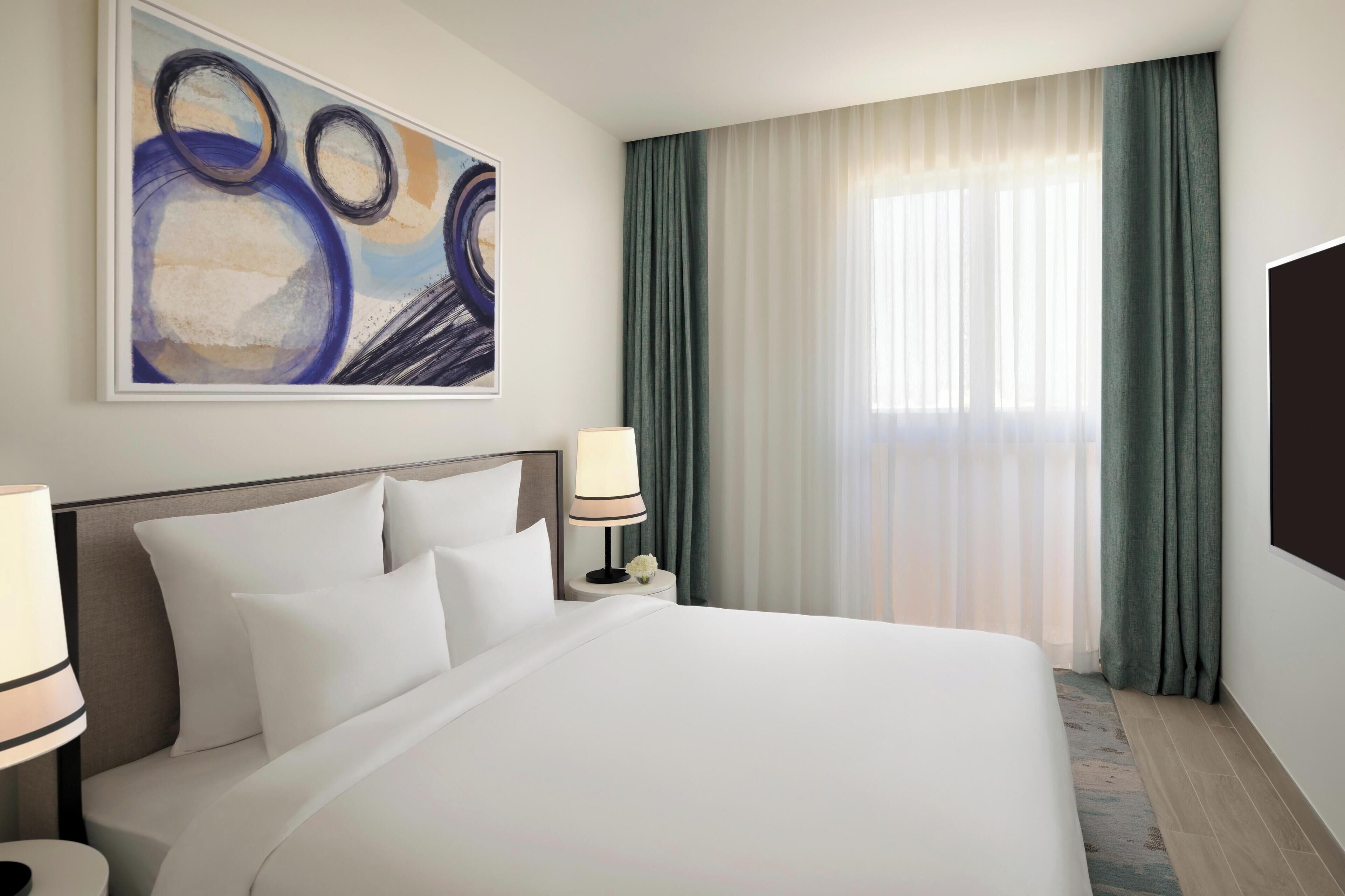 A hotel room with a large bed, a nightstand with a lamp, a wall-mounted TV, and a painting above the bed.