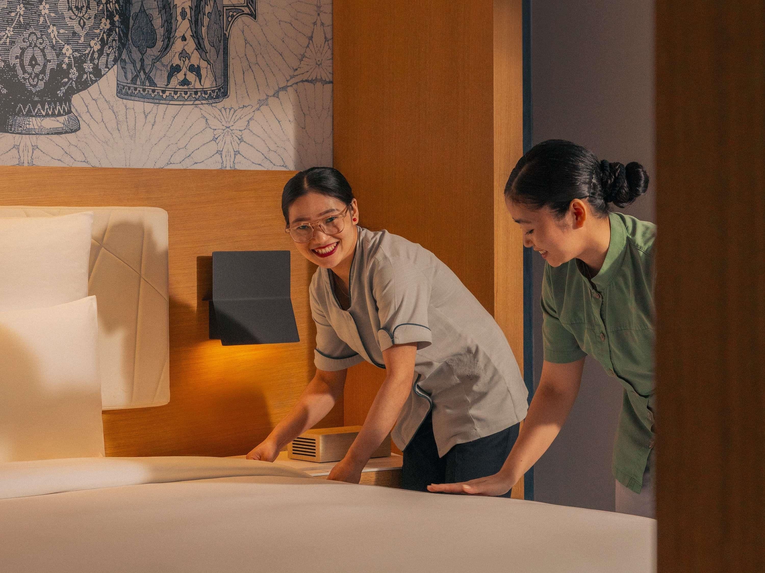 Two individuals arranging a bed in a room with a wooden headboard and a decorative wall hanging.