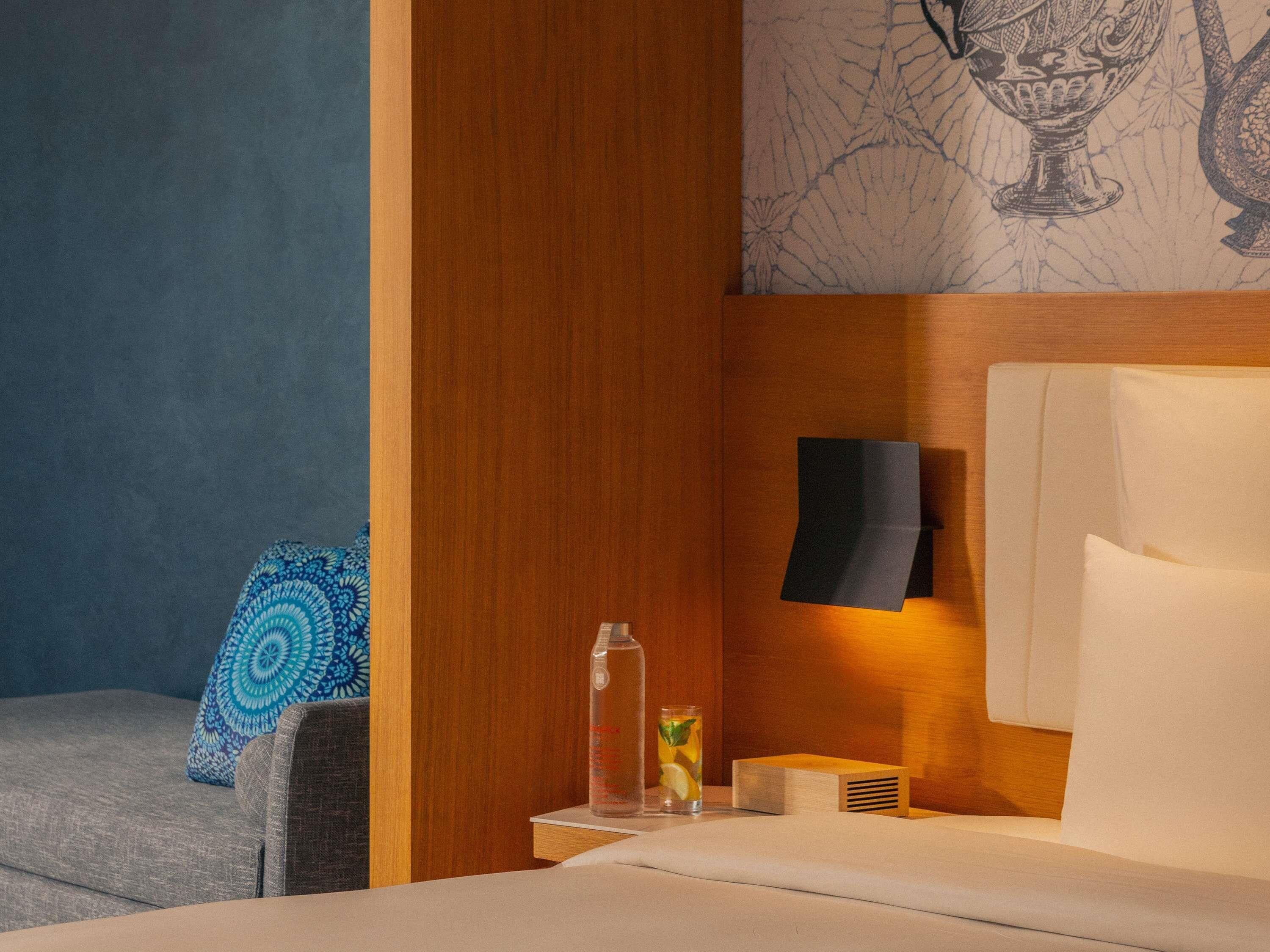 Habitación de hotel con cama, mesita de noche, vaso de agua, almohada decorativa y una pared con diseño ornamental.