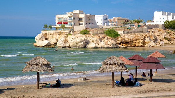 Une plage avec des parasols en chaume, une côte rocheuse et des bâtiments en arrière-plan.