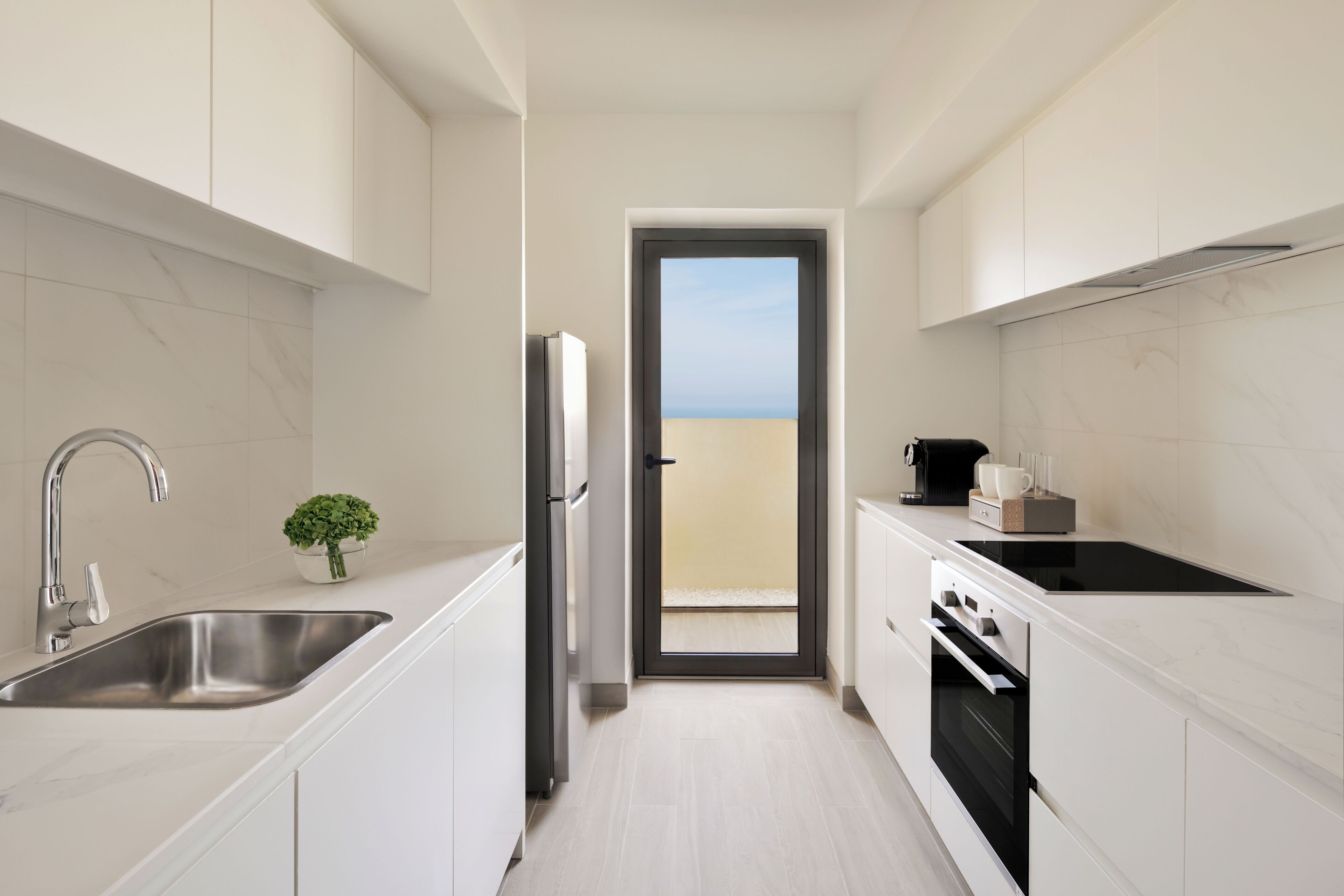 Una cocina moderna con armarios blancos, un lavamanos de acero inoxidable, una estufa negra y vistas al balcón a través de una puerta.