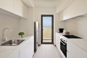 Une cuisine moderne avec des meubles blancs, un évier en acier inoxydable, une cuisinière noire et une vue sur un balcon, accessible par une porte.