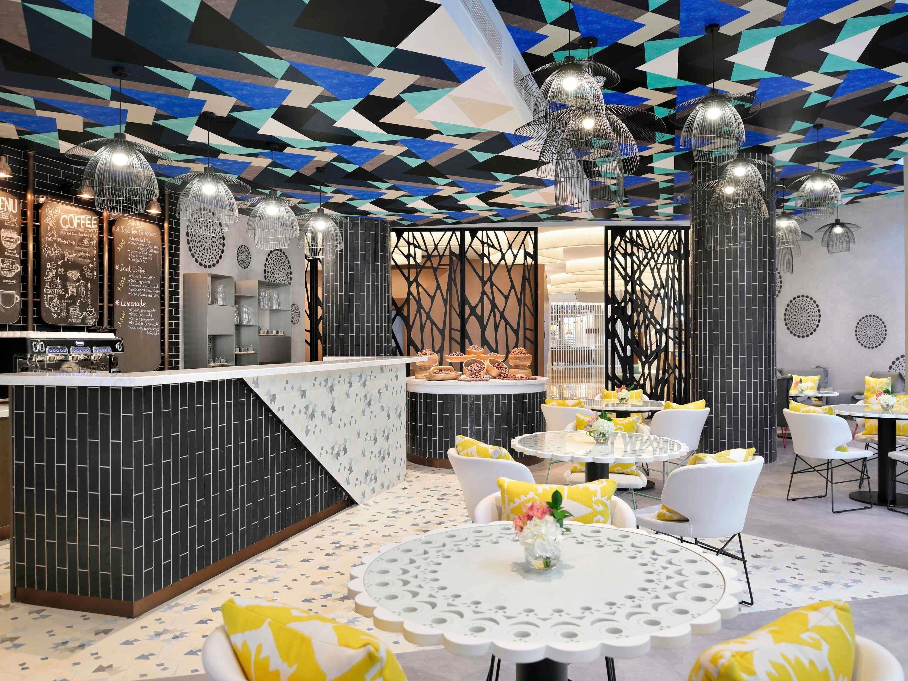 A modern cafe interior with a geometric patterned ceiling, a coffee bar, and round tables with yellow cushions.