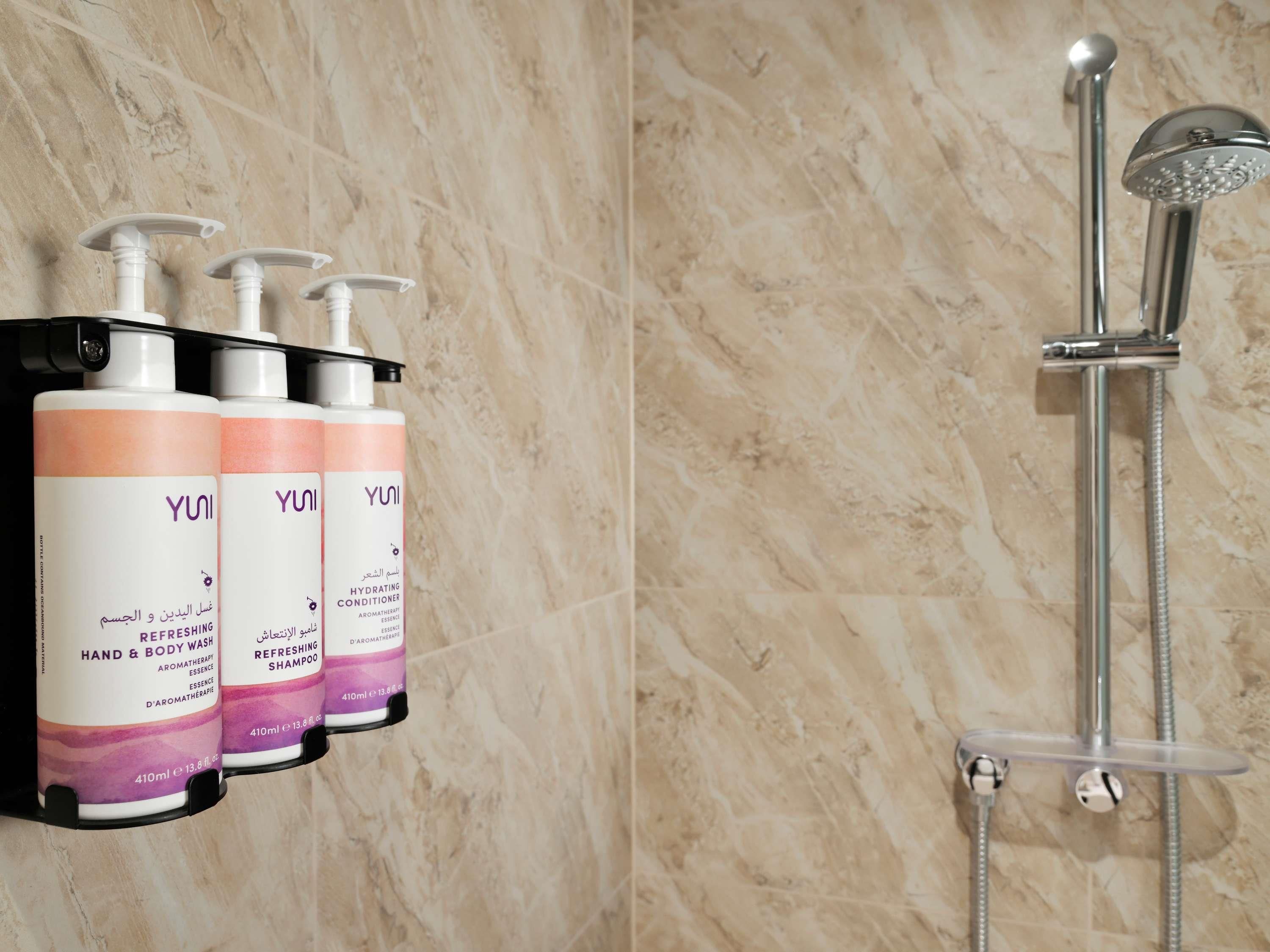 Un baño con pared de mármol, una ducha con cabezal de ducha manual y tres botellas de jabón refrescante para manos y cuerpo YUNI.