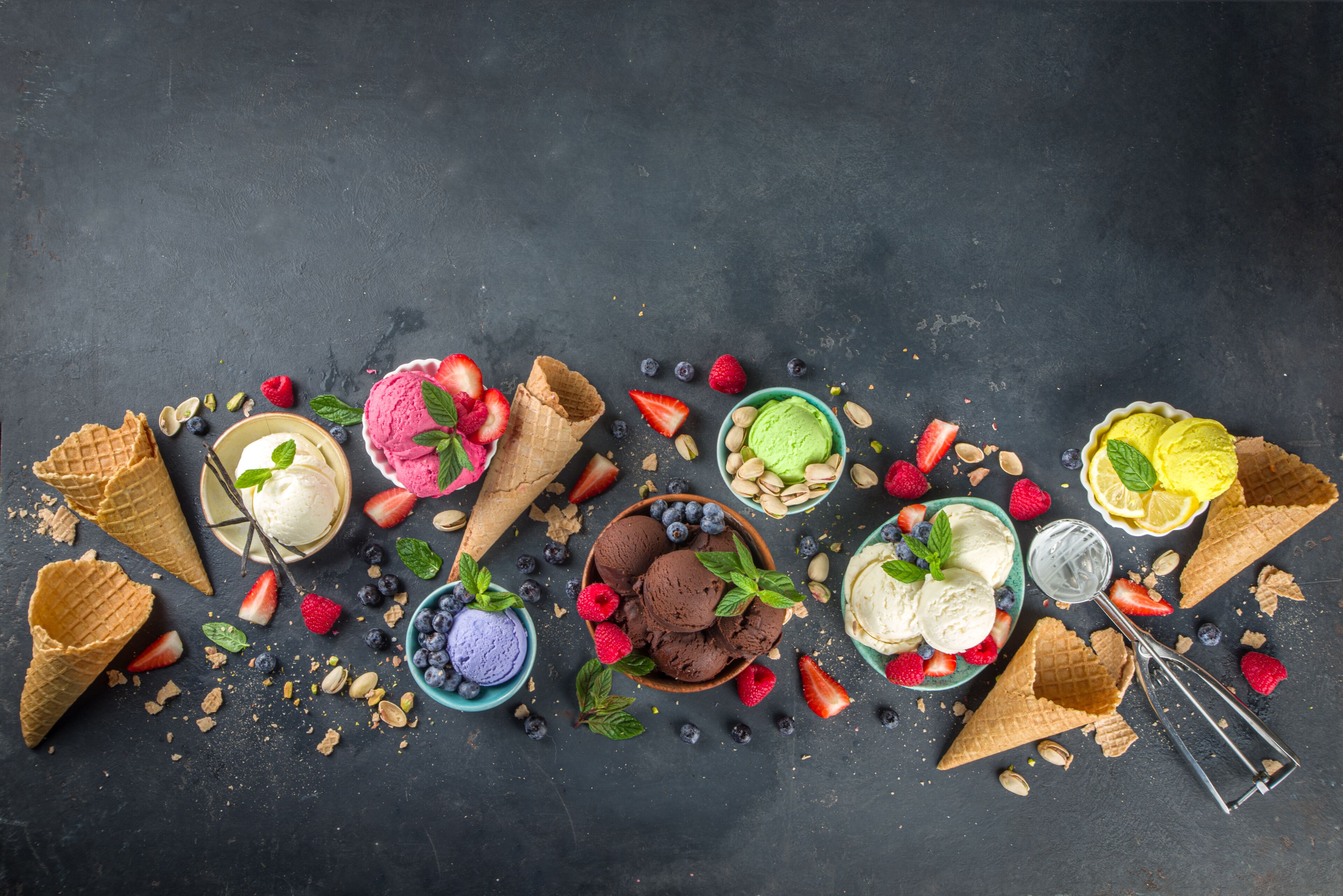 Verschiedene Eissorten auf dunklem Untergrund, dazu Waffelhörnchen und frische Beeren.