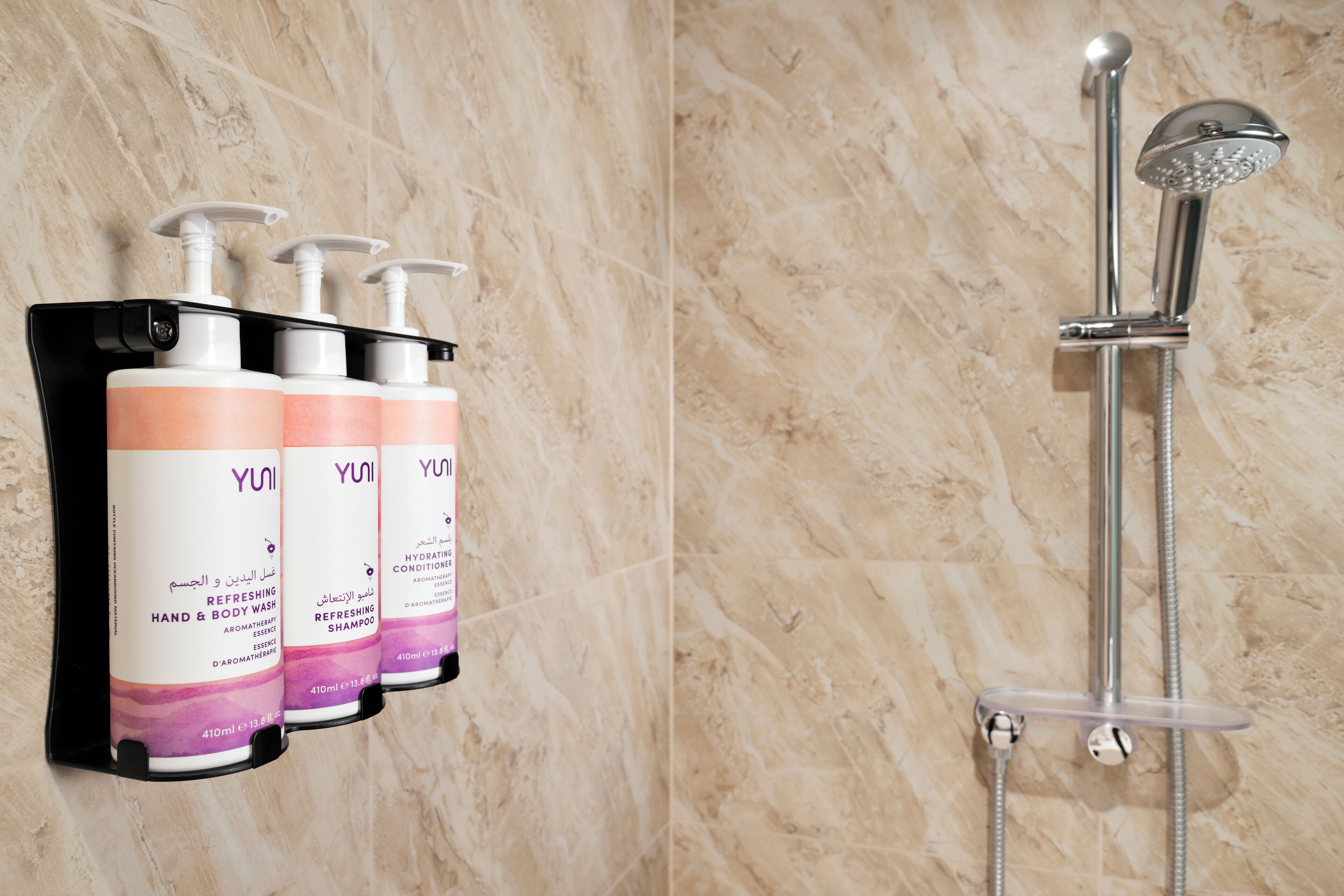 Une salle de bain avec un mur en marbre, un porte-savon noir contenant trois flacons de shampoing rafraîchissant de la marque Yuni, et une douche moderne dotée d’un pommeau de douche à effet pluie.