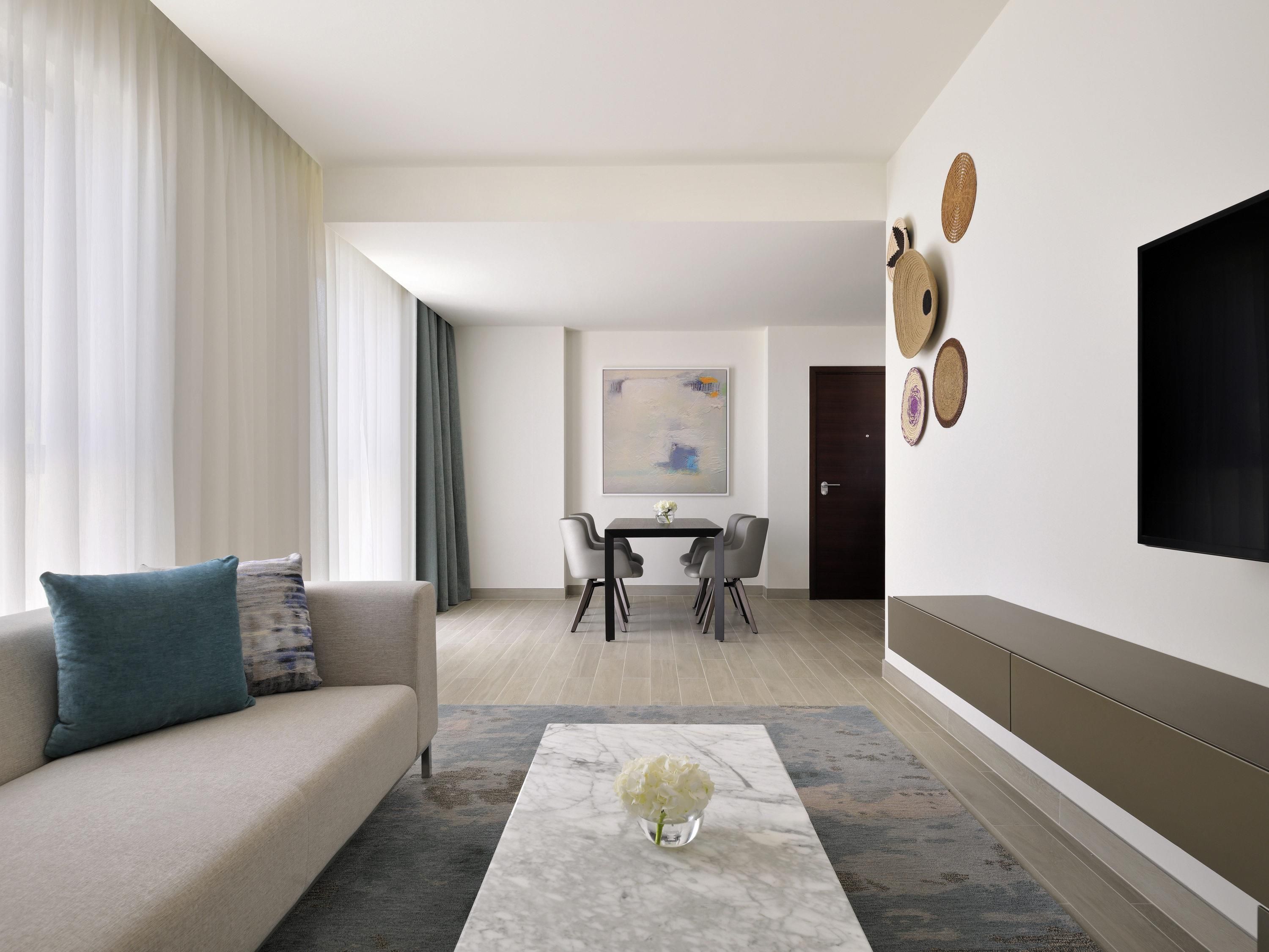 A modern living room with a sofa, a coffee table with a vase, a dining area with chairs, and a flat-screen TV mounted on the wall.