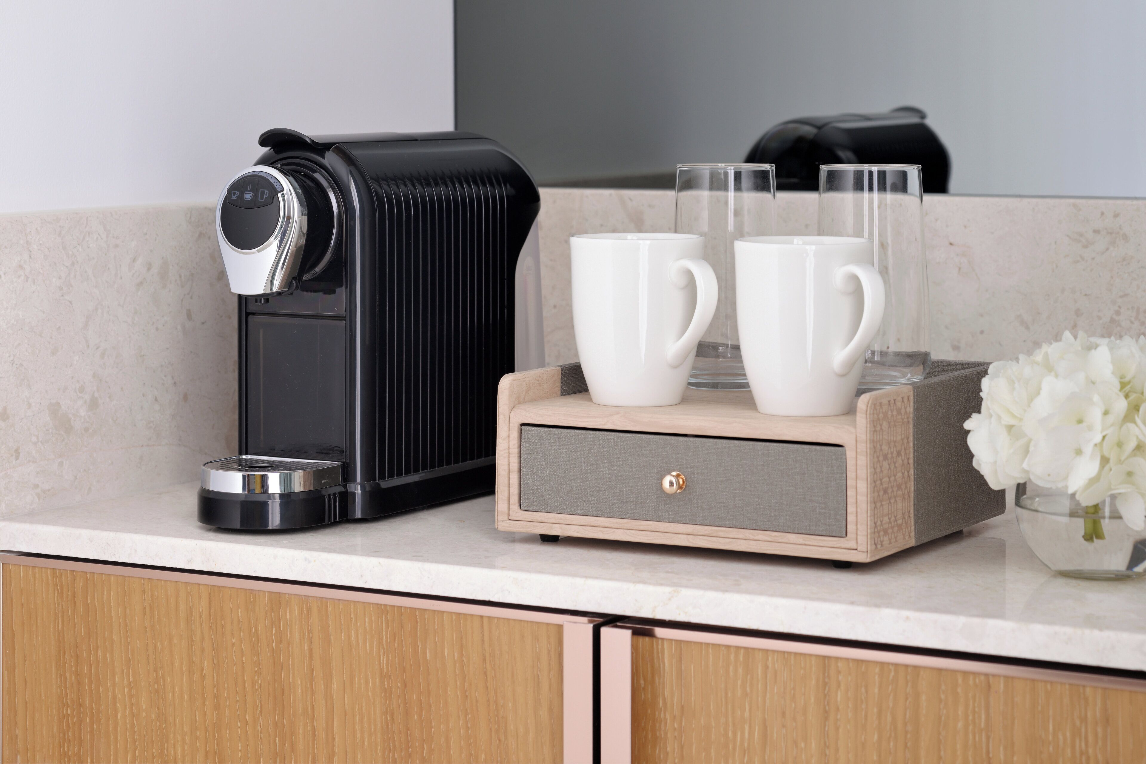 A coffee maker, two white cups, and a glass on a kitchen counter.