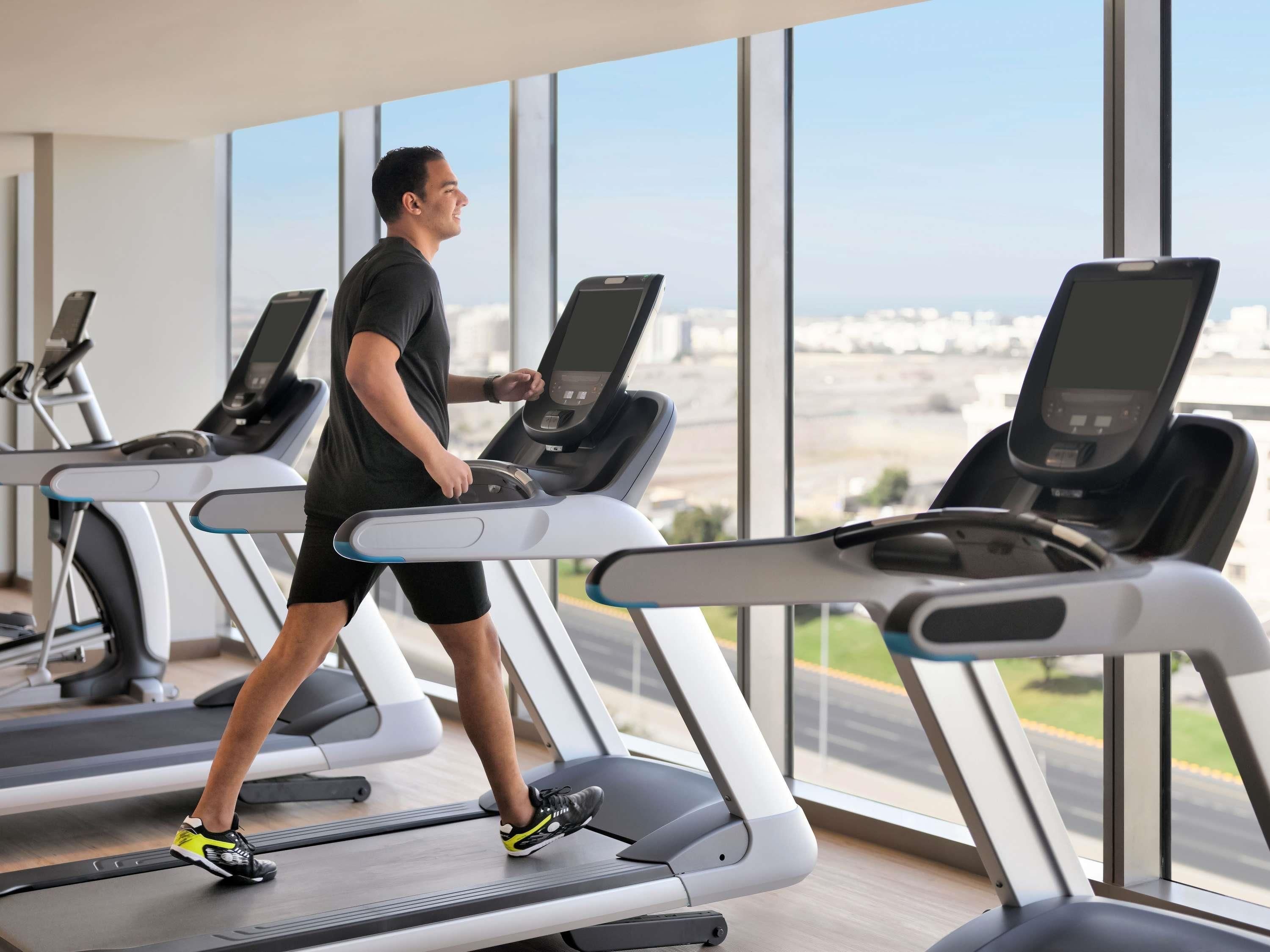 A person walking on a treadmill in a gym with large windows and multiple treadmills.