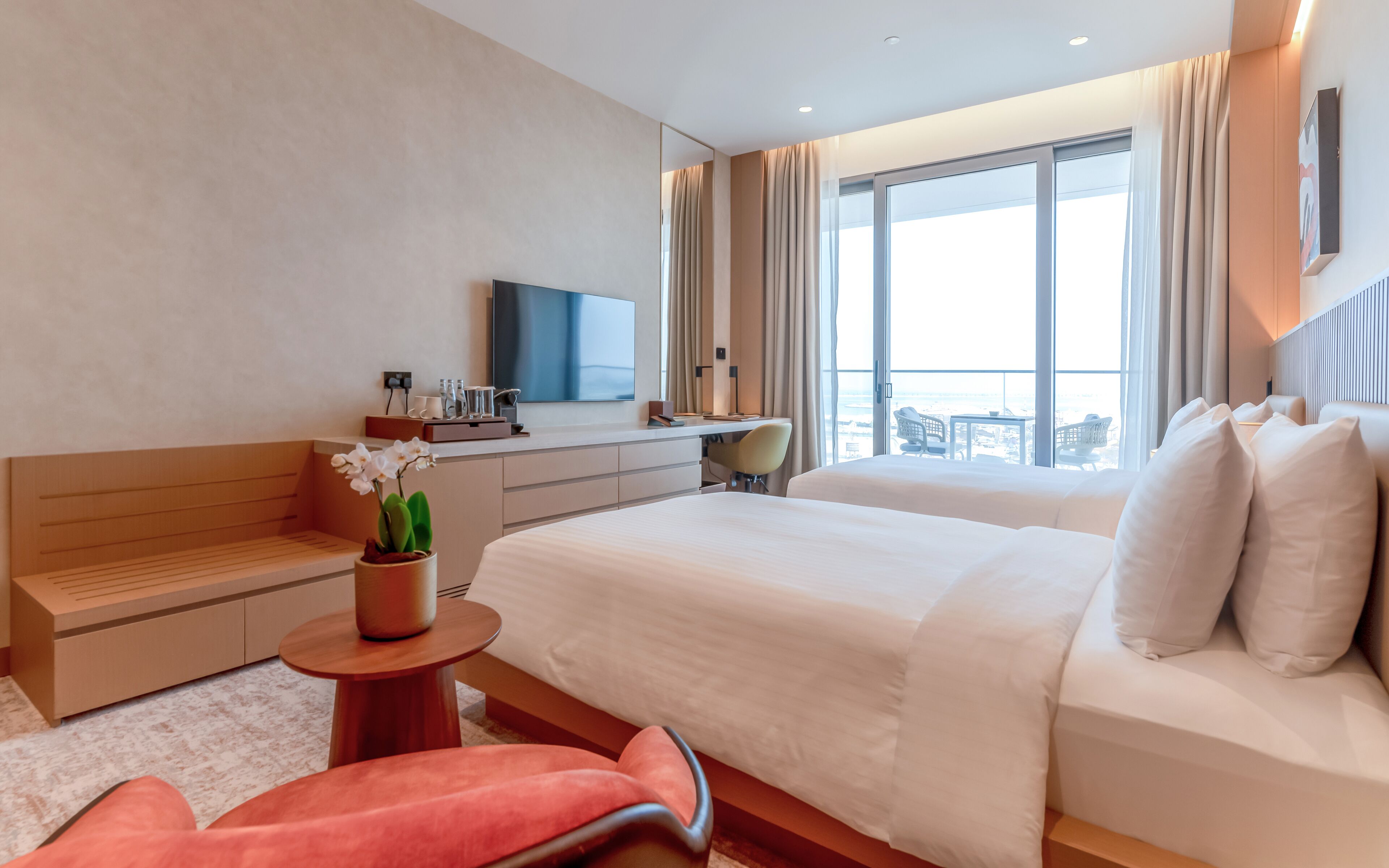 A hotel room with a bed, a desk, a chair, a small table with a plant, and a large window with a view.
