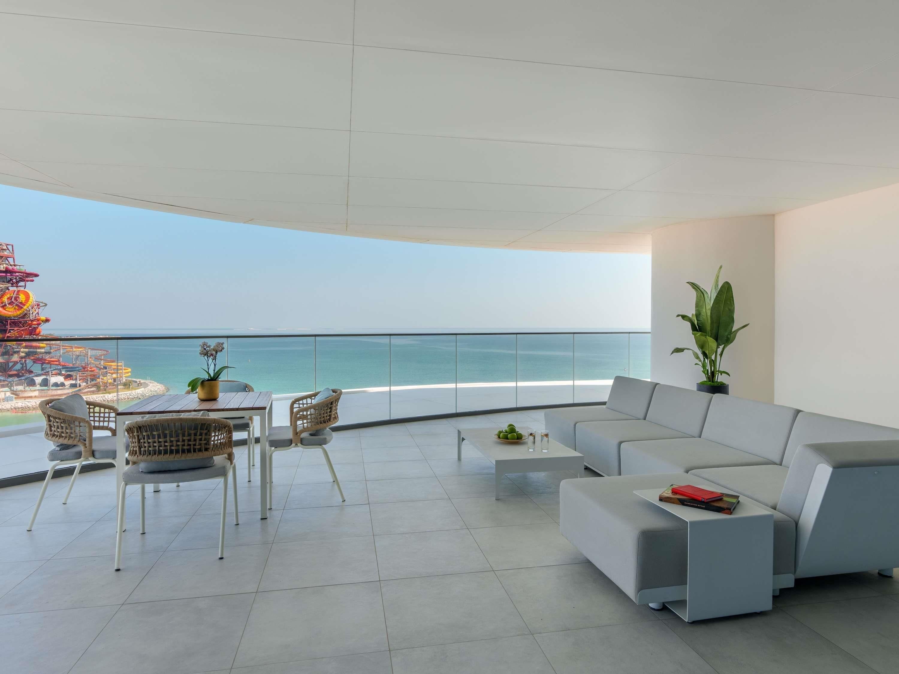 Uma sala de estar moderna com varanda e vista para o mar, com um sofá, cadeiras, uma mesinha e um vaso de planta.