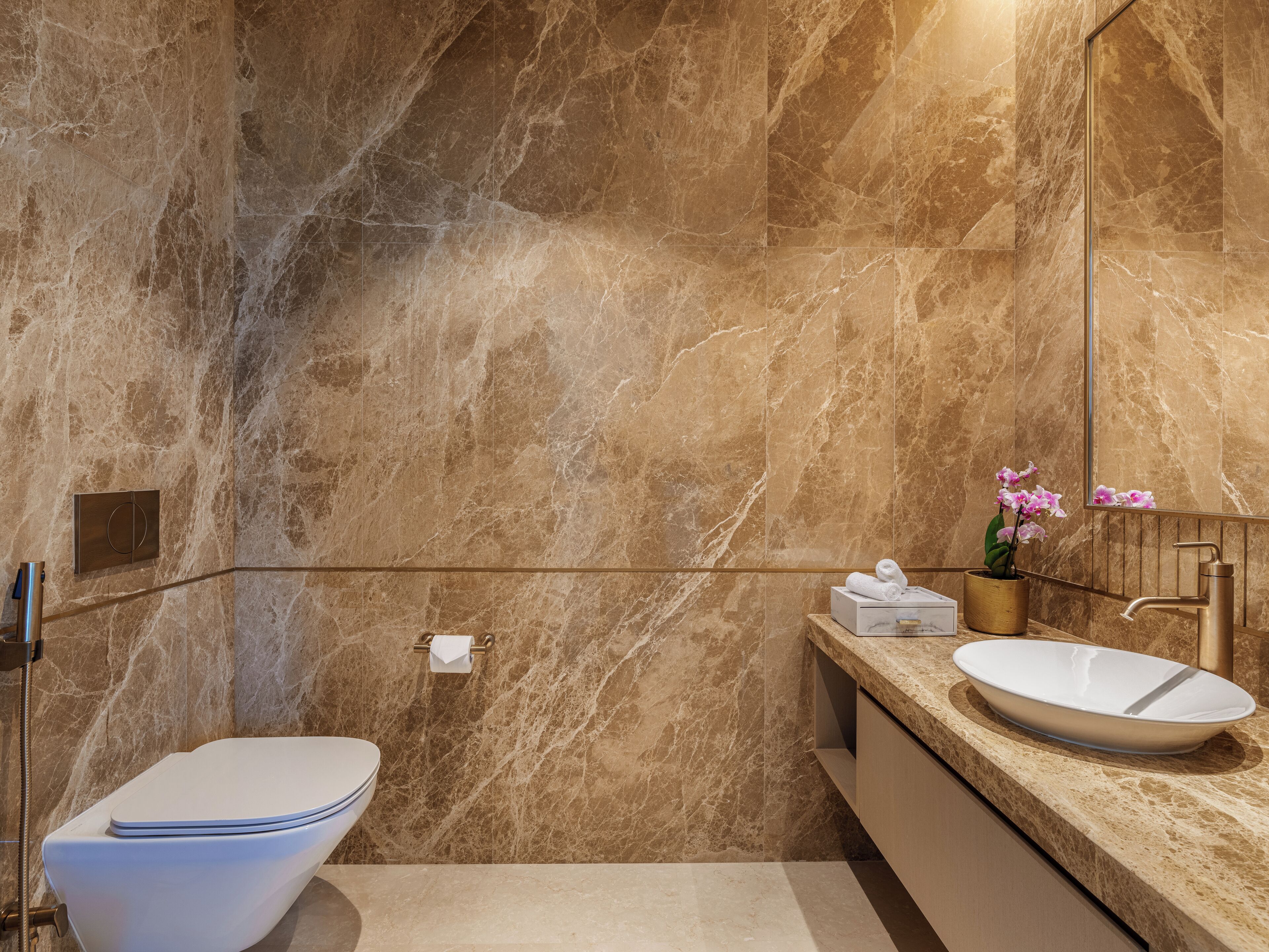 Un baño moderno con pared de mármol, inodoro blanco, lavabo con grifo dorado y espejo.