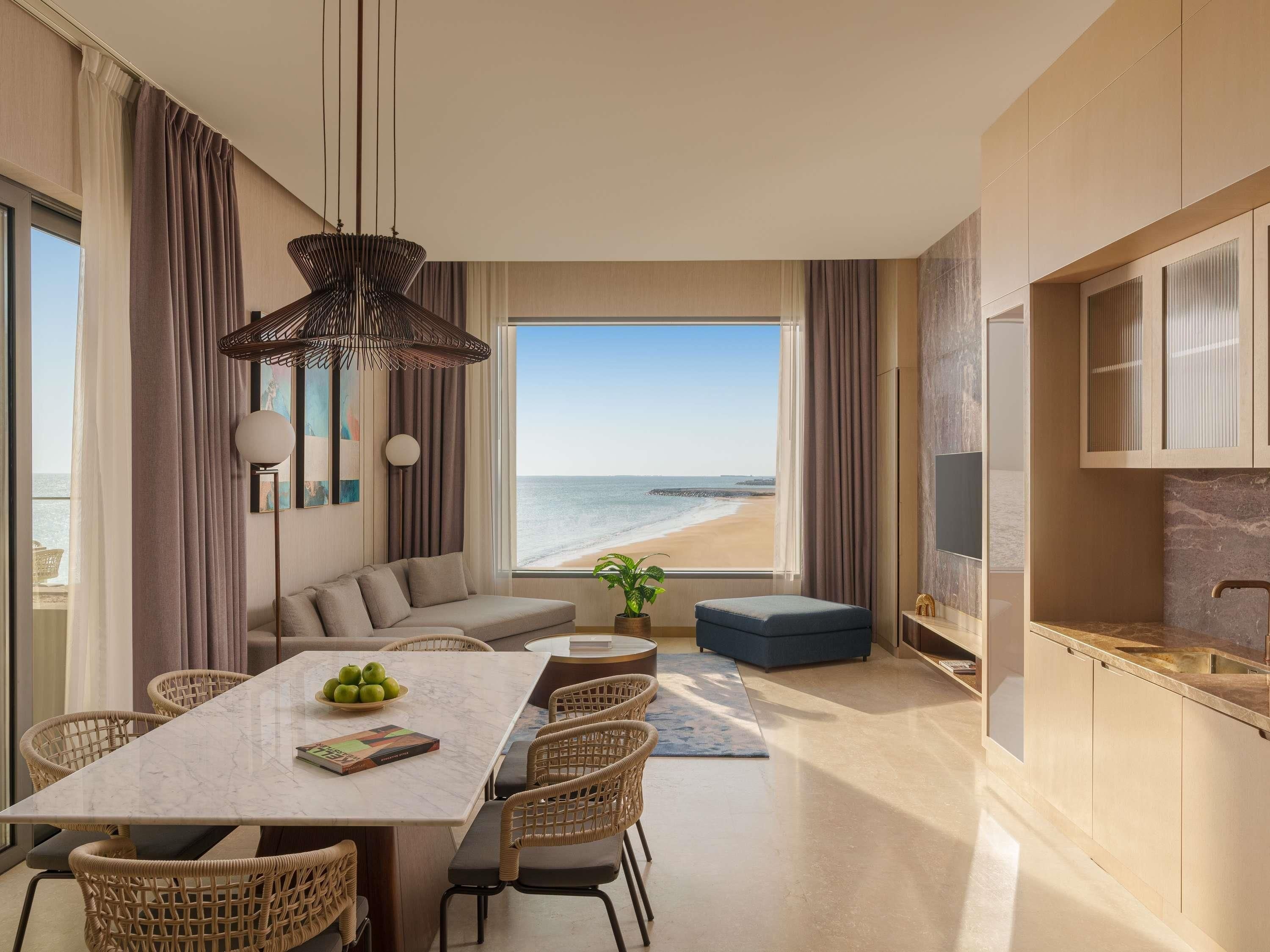 Una sala moderna con un ventanal que da a una playa, un comedor con mesa de mármol y una kitchenette con televisor.