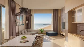 Una sala moderna con un ventanal que da a una playa, un comedor con mesa de mármol y una kitchenette con televisor.