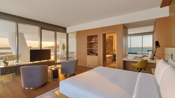 A modern hotel room with a large bed, a desk with a chair, a TV, and a balcony with a view of the city.