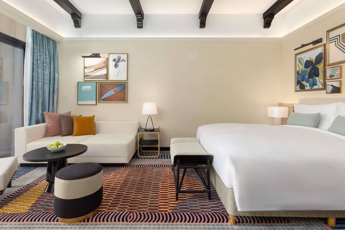 Een hotelkamer met een bed, een bank, een klein tafeltje en een stoel. Er hangen kunstwerken aan de muur en er is een raam met gordijnen.
