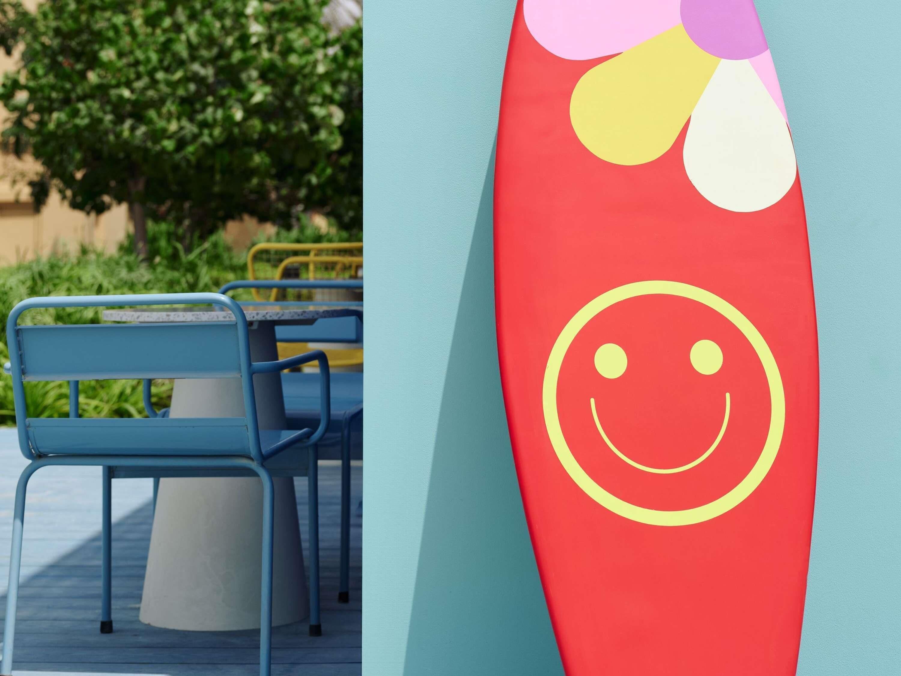 A blue metal chair and a red surfboard with a yellow smiley face.