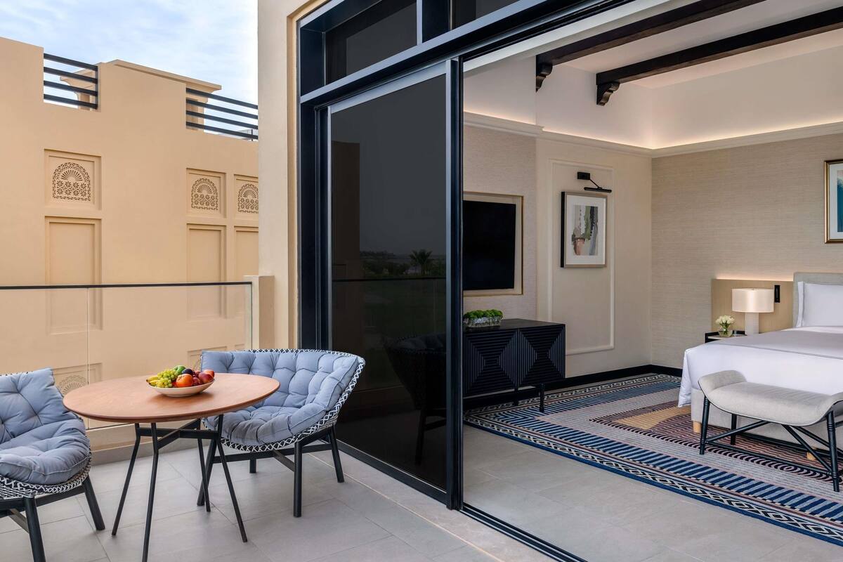 Een moderne hotelkamer met balkon, een zithoek met een ronde tafel en stoelen, en een kamerkleed met patroon.