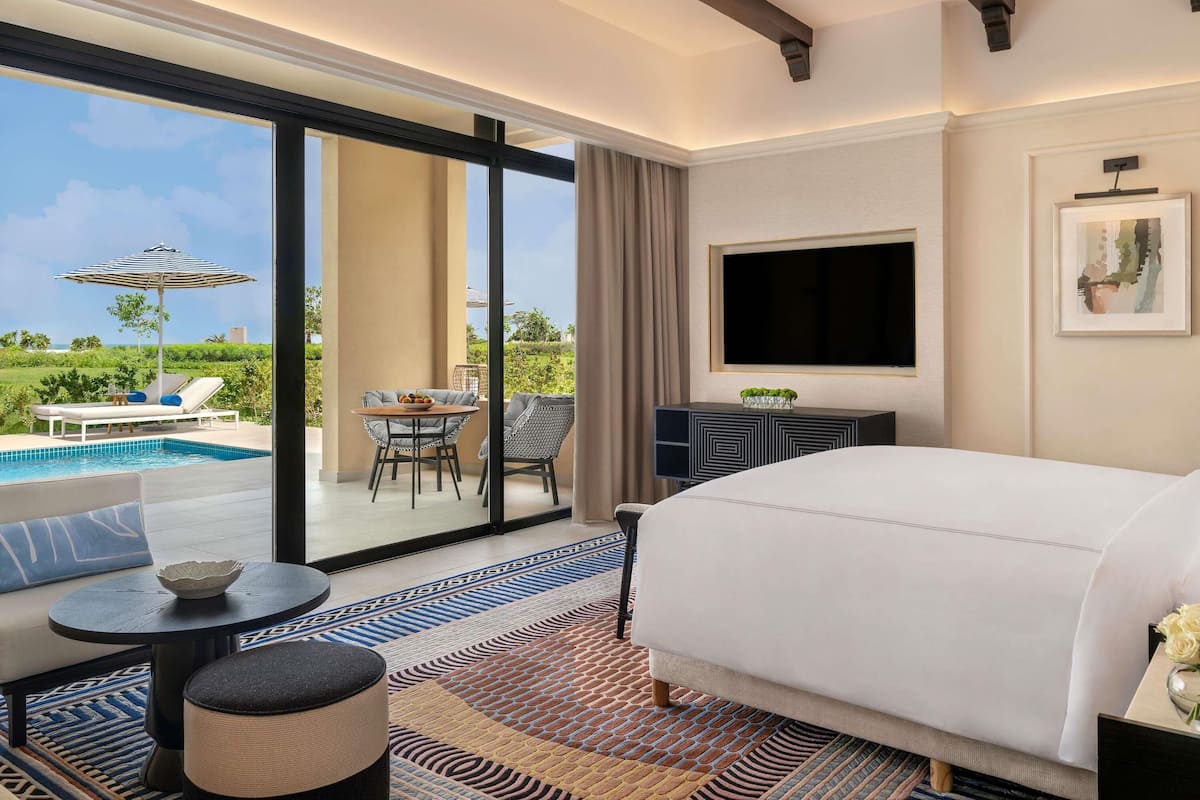 Een hotelkamer met een groot bed, een televisie, een zithoek met een tafel en stoelen, en uitzicht op een zwembad en groen buiten.