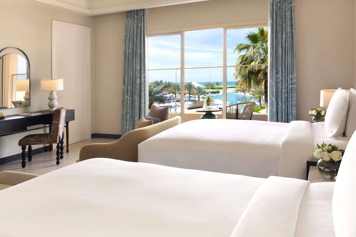 Hotelkamer met twee bedden, een bureau, een stoel, een groot raam met uitzicht op het zwembad en palmbomen, en een spiegel.